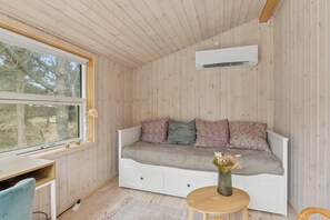 Et hyggeligt rum med træpaneler, en hvid sofa med blomstermønstrede puder, et lille bord med en vase og et vindue med udsigt til træer.