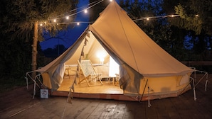 Ein großes, beige Zelt mit Holzboden und einem Bett im Inneren, umgeben von Bäumen und Lichterketten.