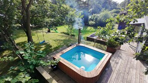 Un jacuzzi rodeado por una terraza de madera, una hamaca y un césped con árboles.
