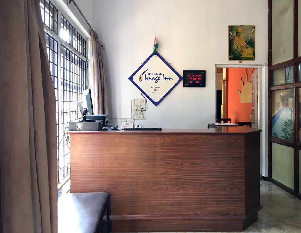 A hotel reception desk with a wooden finish, a sign for Hotel Grand Image Inn, a digital clock, and a framed picture on the wall.