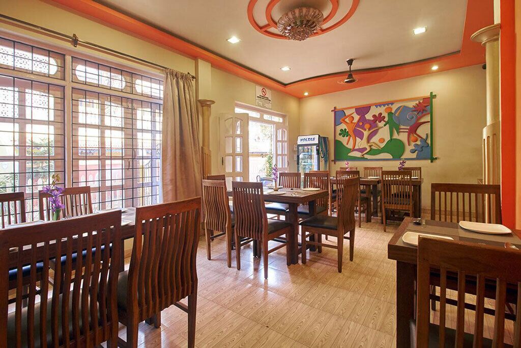 A dining area with wooden tables and chairs, a large window with curtains, and a colorful abstract painting on the wall.