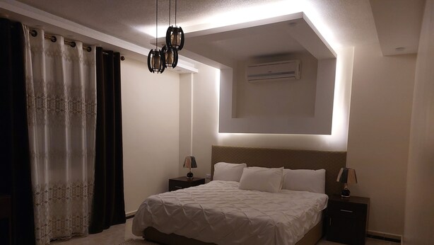 A bedroom with a bed, two bedside tables, a hanging light fixture, an air conditioning unit, and a window with curtains. - Petra Guests Home  (Wadi Musa)
