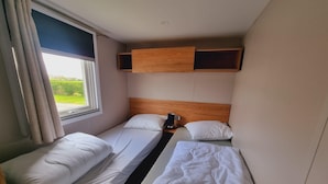 A small, compact room with two single beds, a window with curtains, and a wooden shelf above the beds. - Chalet in Belgium with Garden Terrace (Blankenberge)