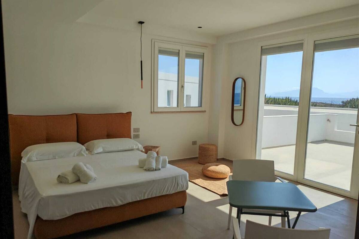 A modern bedroom with a large bed, a small table, and a view of the sea through a sliding door.