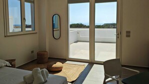A hotel room with a bed, a chair, a small round ottoman, a mirror, and a sliding glass door leading to a balcony with a view of the sea and buildings. - Dimora Cala Graziosa (Favignana)