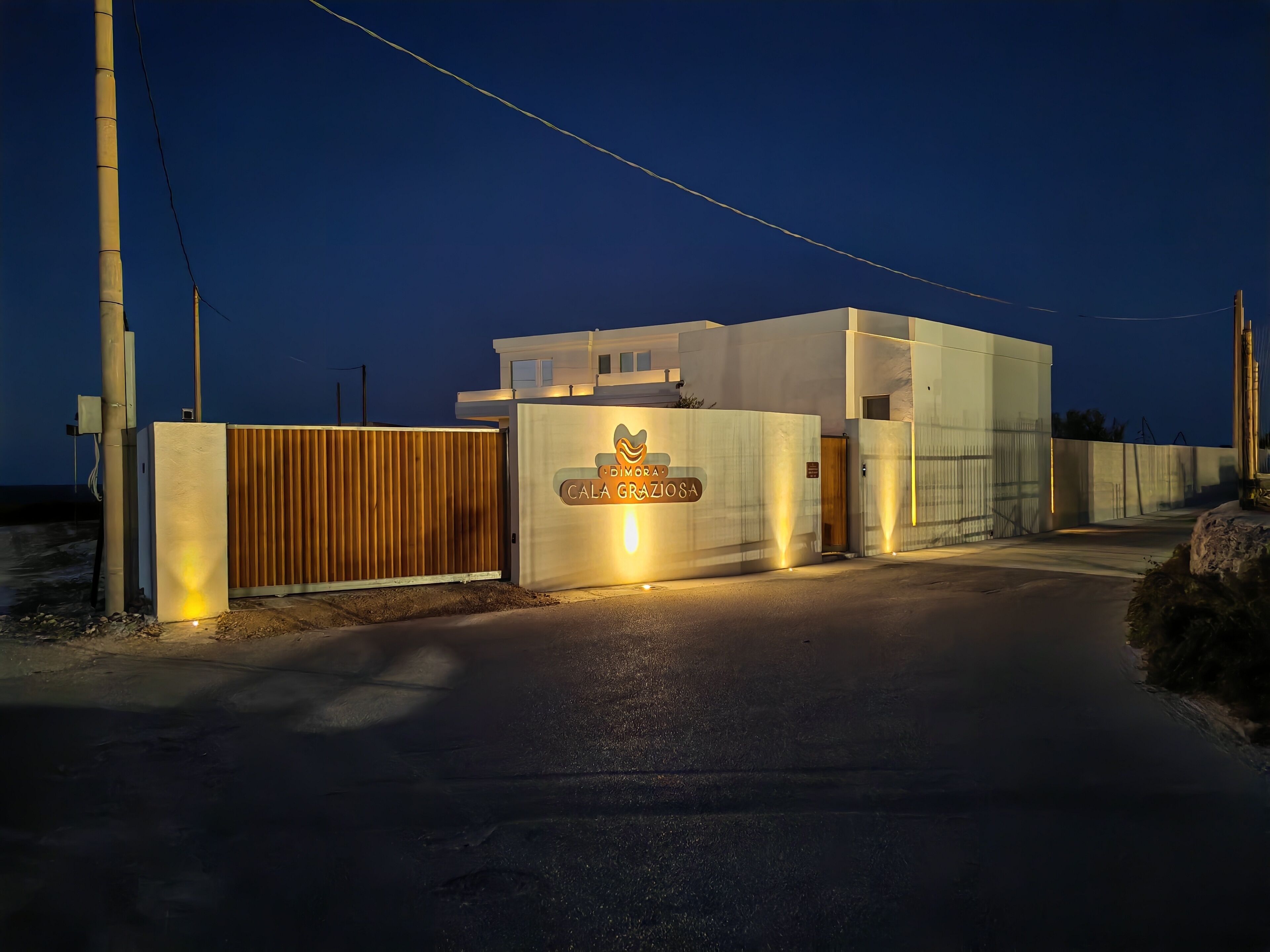 A modern building with a gated entrance, a sign reading 'Dimora Cala Grazia,' and a clear sky at dusk.