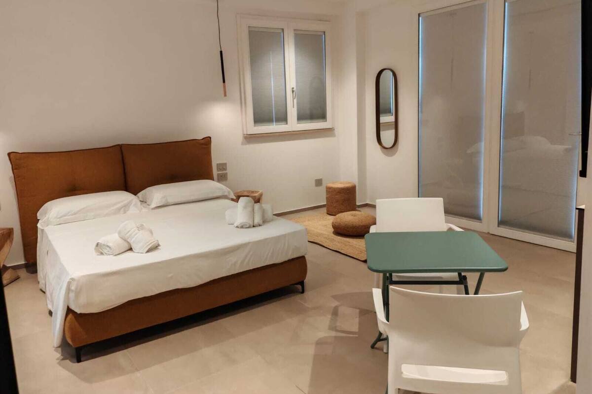A modern bedroom with a large bed, a small table, a chair, and a sliding glass door.