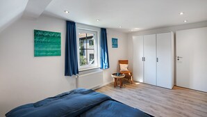 Ein Schlafzimmer mit Bett, einem Fenster mit Vorhängen, einem Sessel, einem Kleiderschrank und einer Heizung.