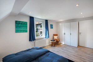 Un dormitorio con cama, ventana con cortinas, una silla, armario y radiador.