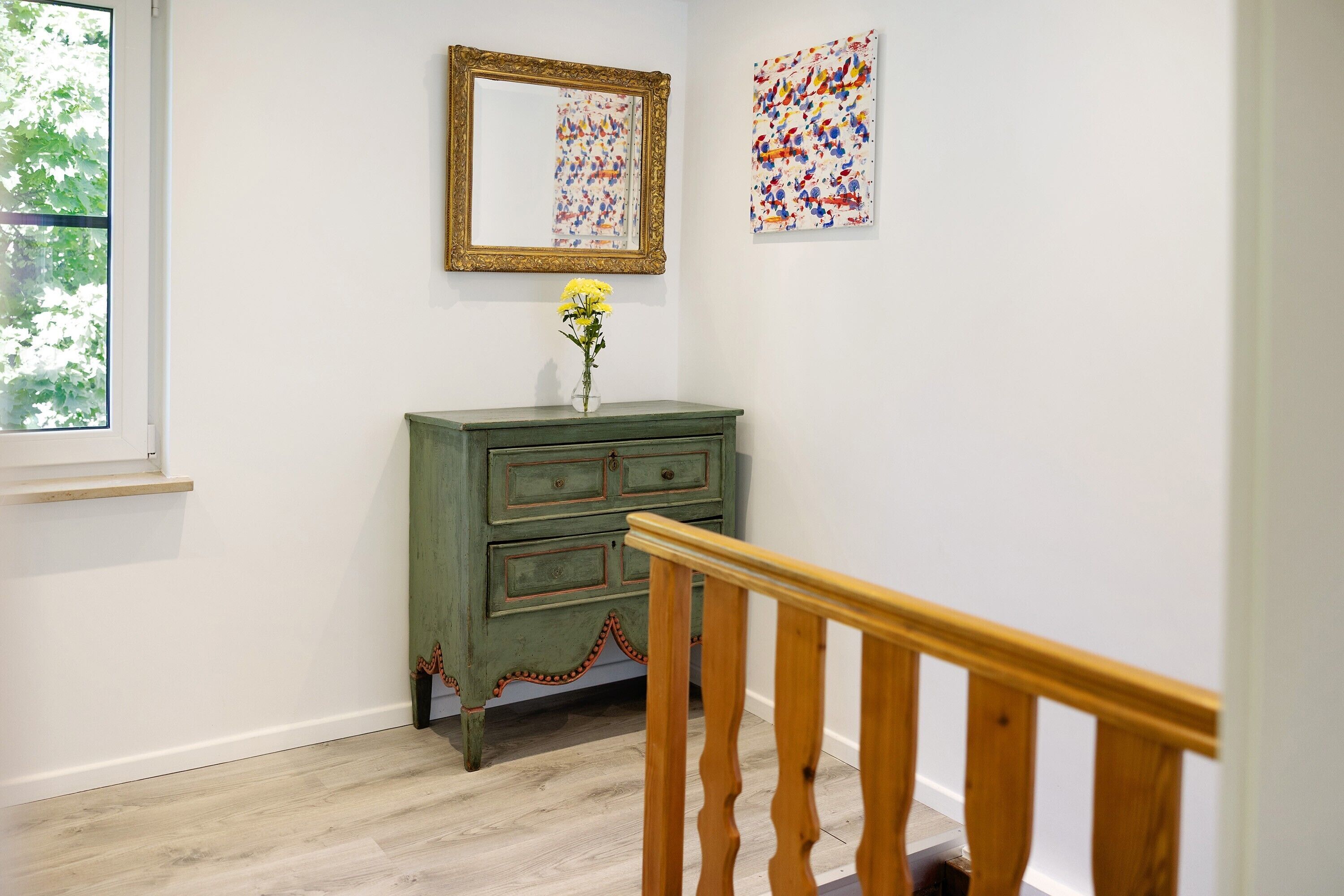 Ein Zimmer mit einer Holztreppe, einem grünen Kommode mit Schubladen, einem Spiegel und zwei Kunstwerken an der Wand.