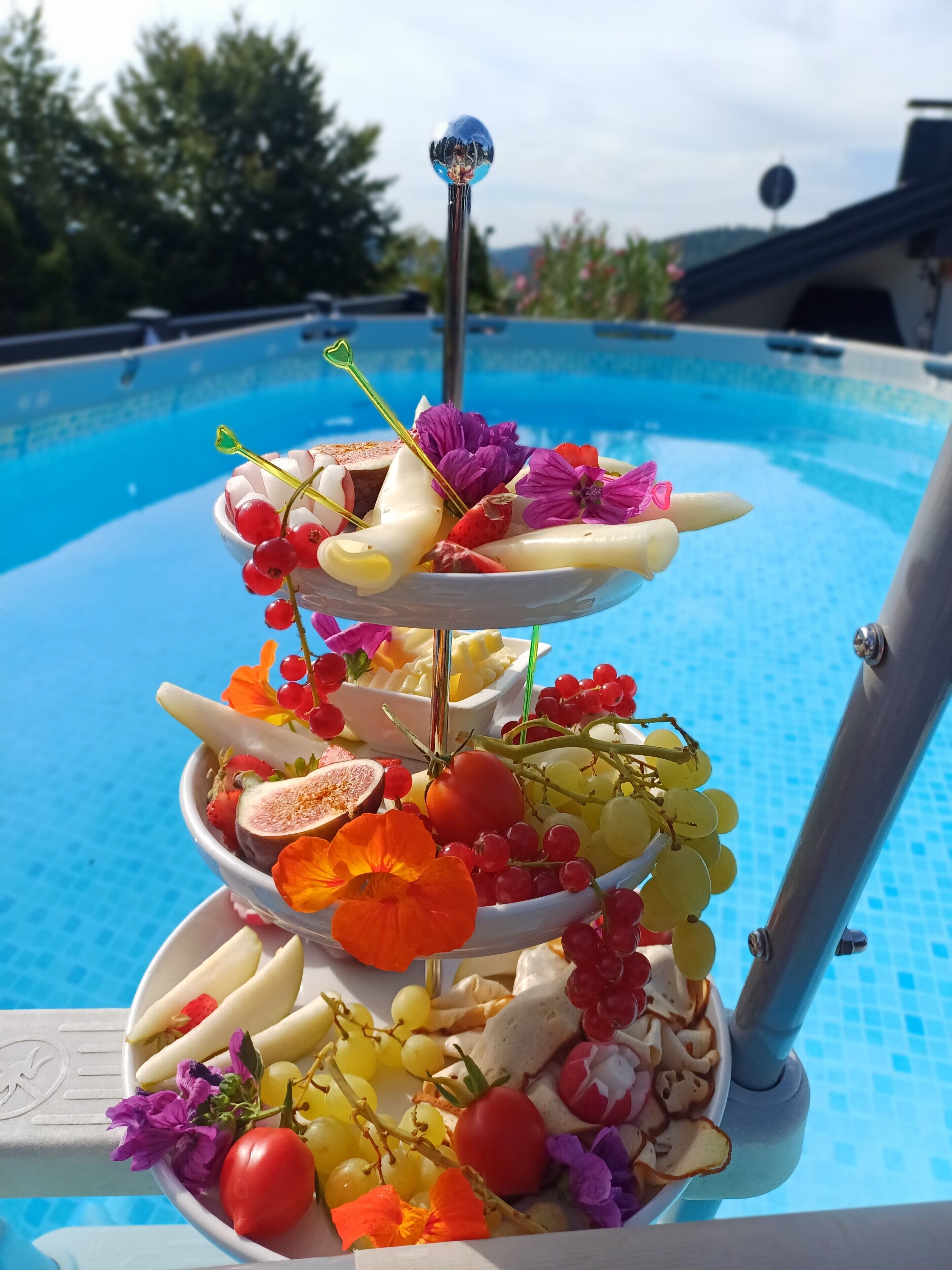 Una bandeja de frutas y quesos apilada junto a una piscina.