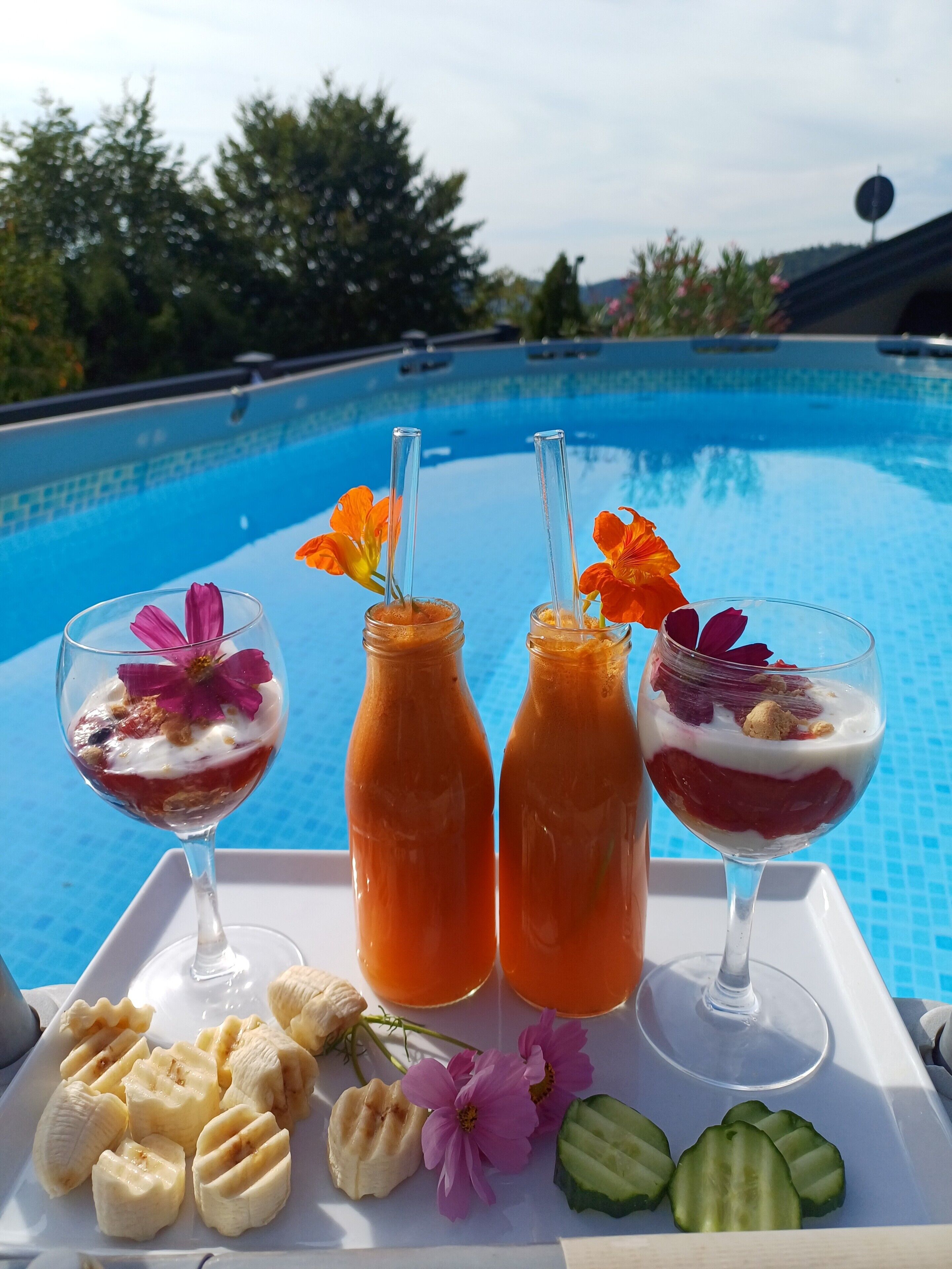 Escena junto a la piscina con una bandeja de comida y bebidas, que incluye rodajas de fruta y dos vasos con pajitas.