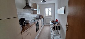 A narrow kitchen with a window, white tiled backsplash, wooden cabinets, and a small dining area with a chair.