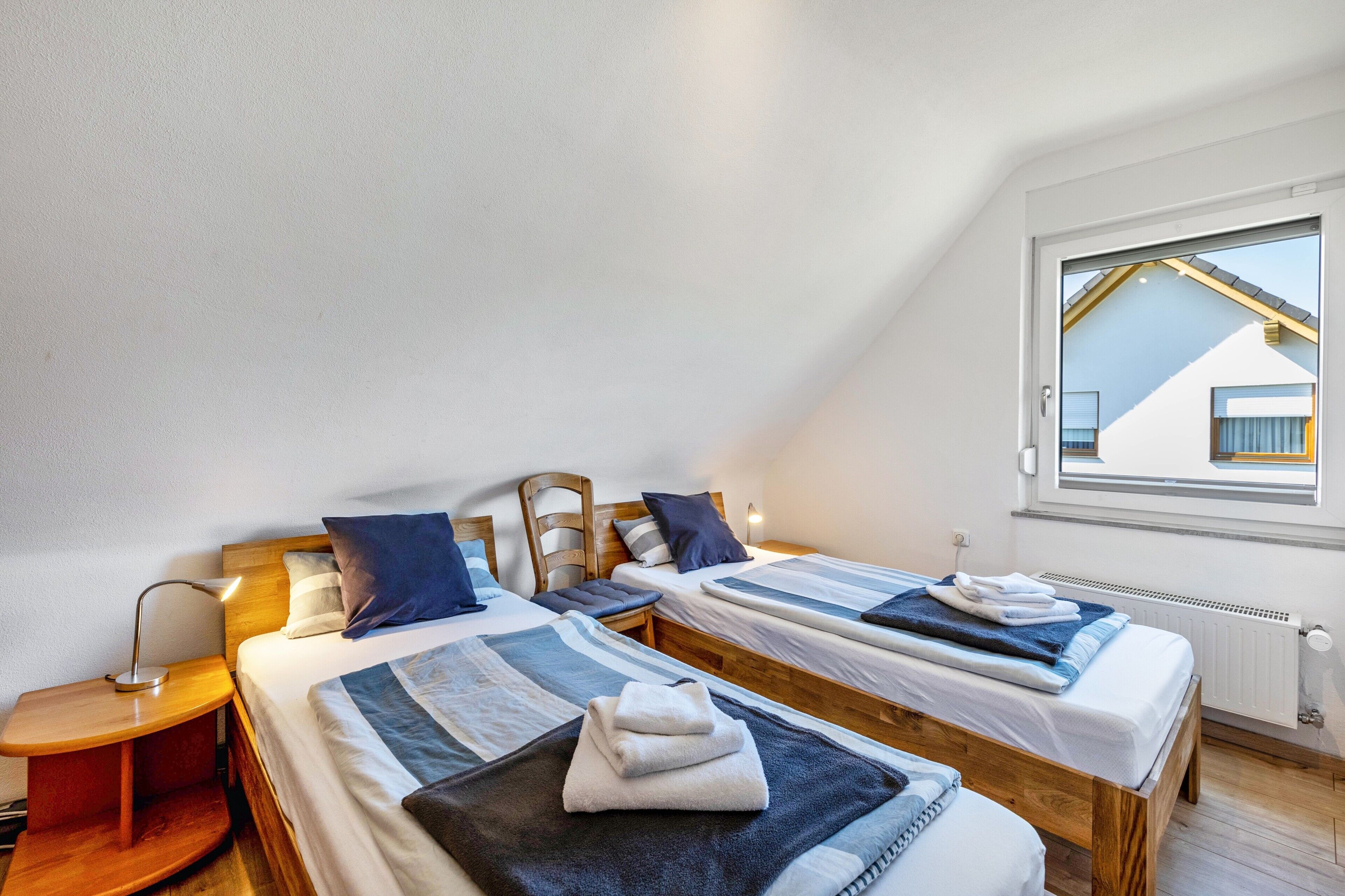 Un dormitorio con dos camas, una mesita de noche, una lámpara y una ventana con vistas a un edificio.