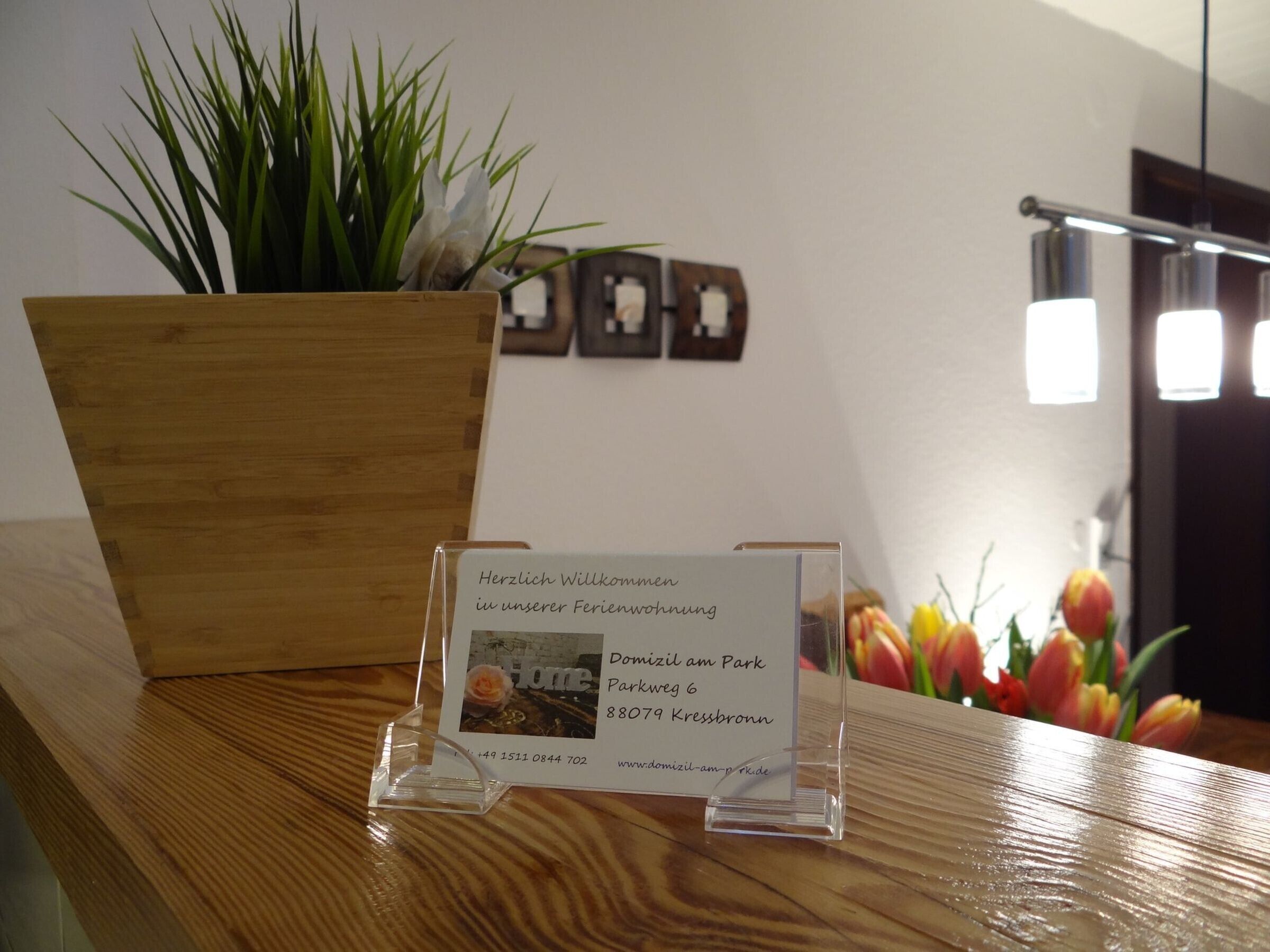 Una recepción con un cartel de bienvenida, una jardinera de madera con plantas verdes y una pared con cuadros enmarcados.