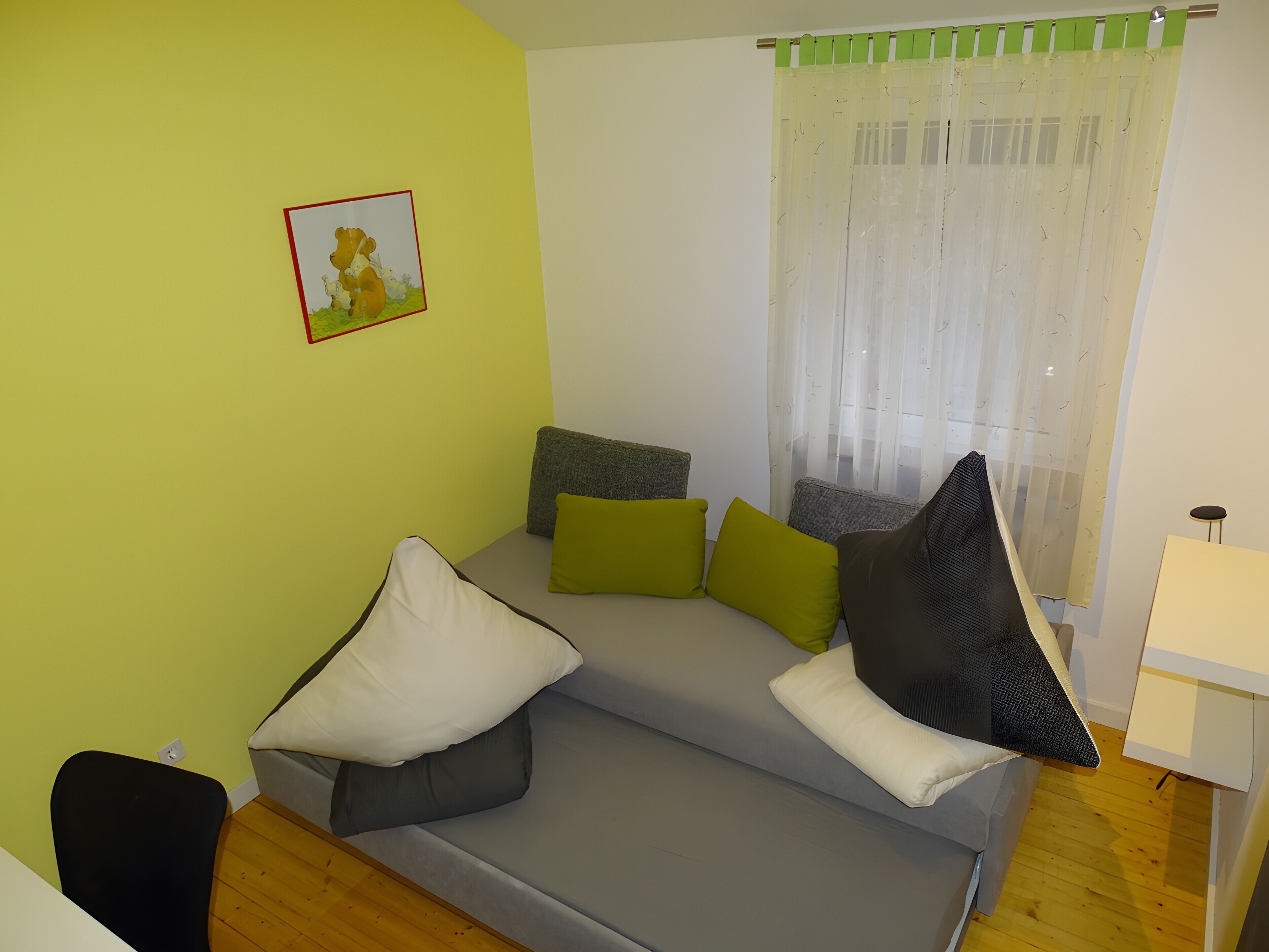 Une pièce avec un canapé gris, des coussins verts et blancs, une petite table et une fenêtre ornée de rideaux légers.