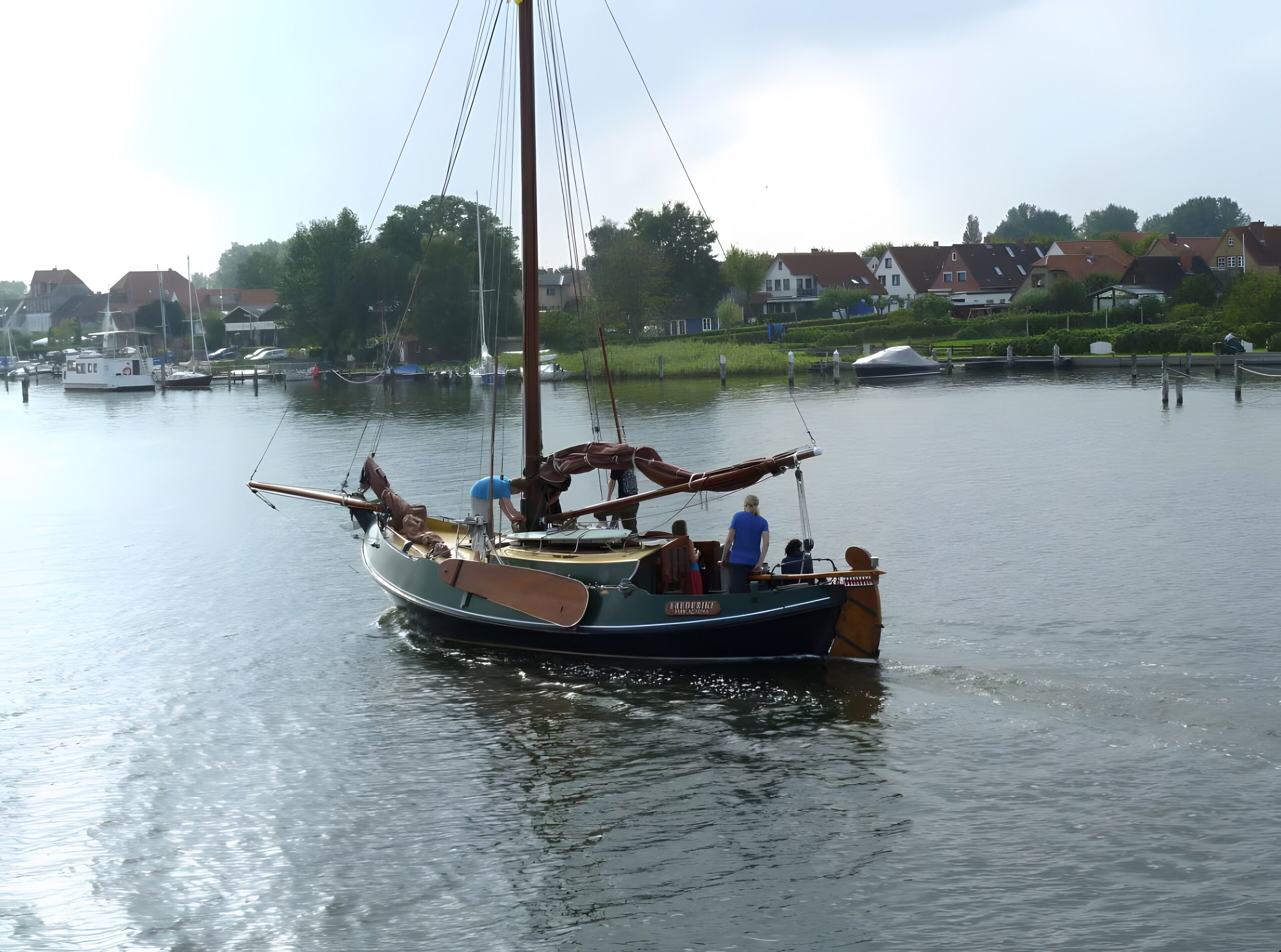 A sailboat with a cabin and a mast, docked at a calm water body with houses in the background.