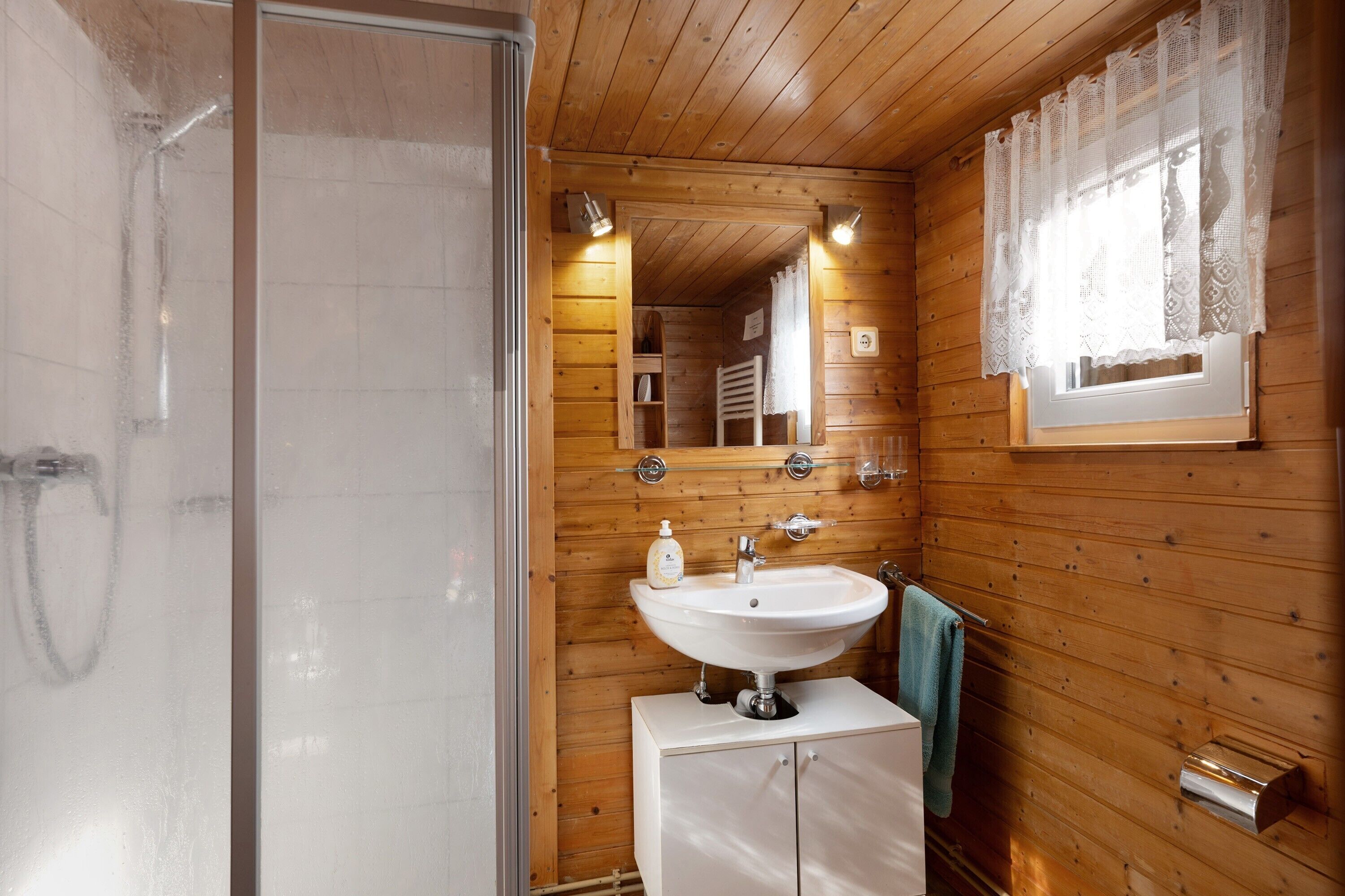 Baño de cabaña de madera con ducha, lavabo y espejo.