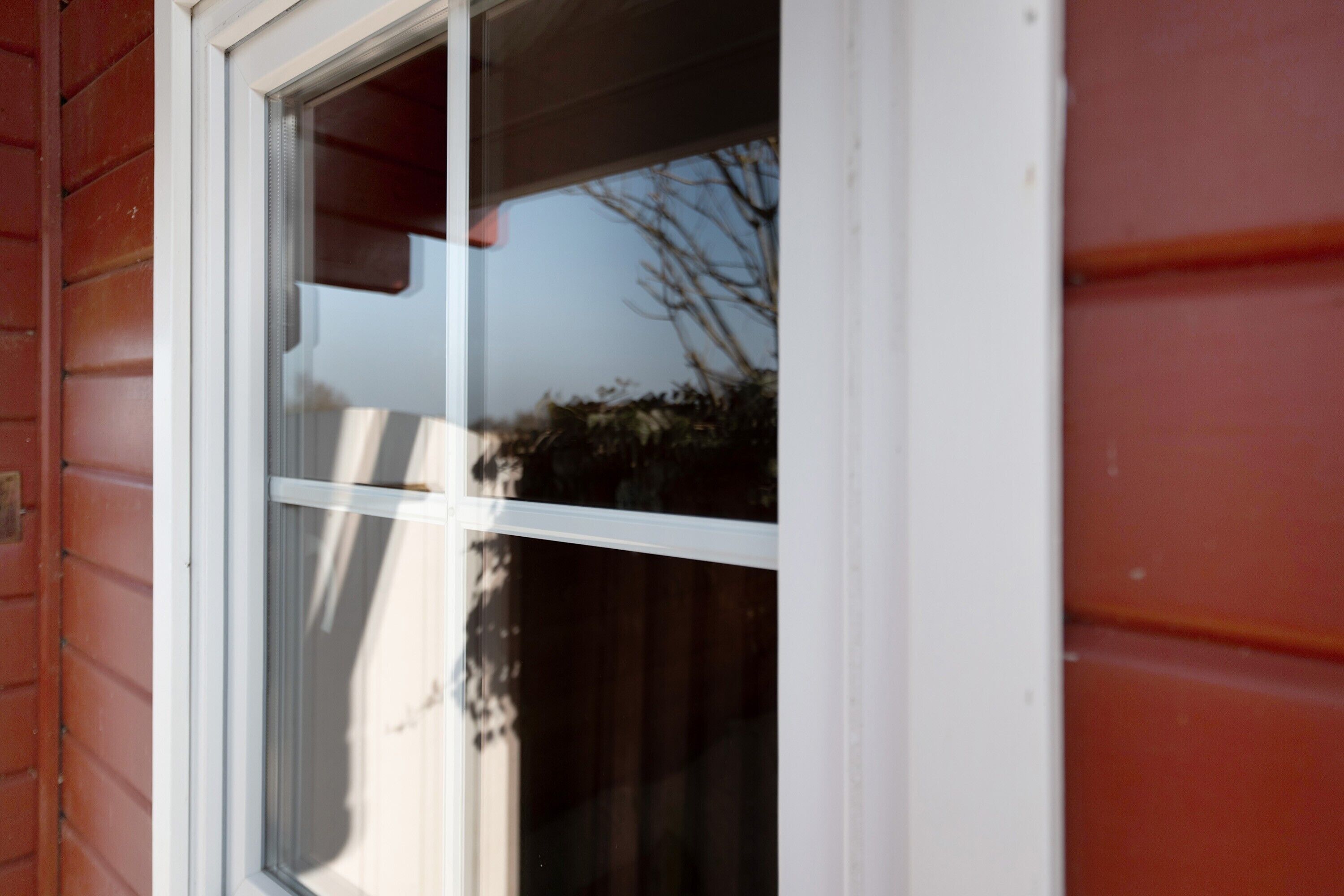 Ein Fenster mit weißem Rahmen, eine Baumreflexion und ein Blick auf ein rotes Gebäude.