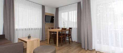 A modern room with a wooden dining table, chairs, a TV, and large windows with sheer curtains.
