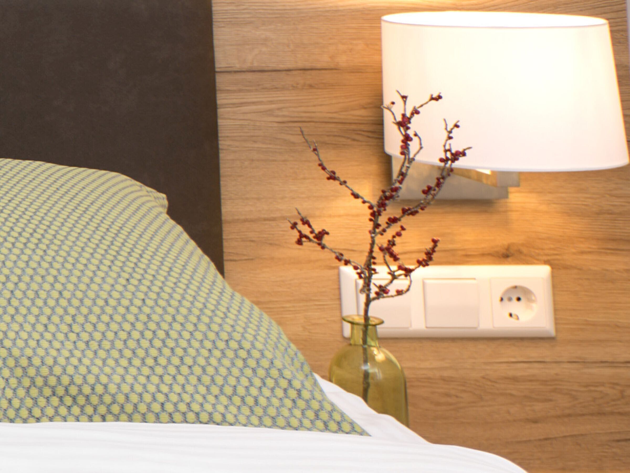 A bed with patterned bedding, a vase with branches, a wall-mounted lamp, and a power outlet.