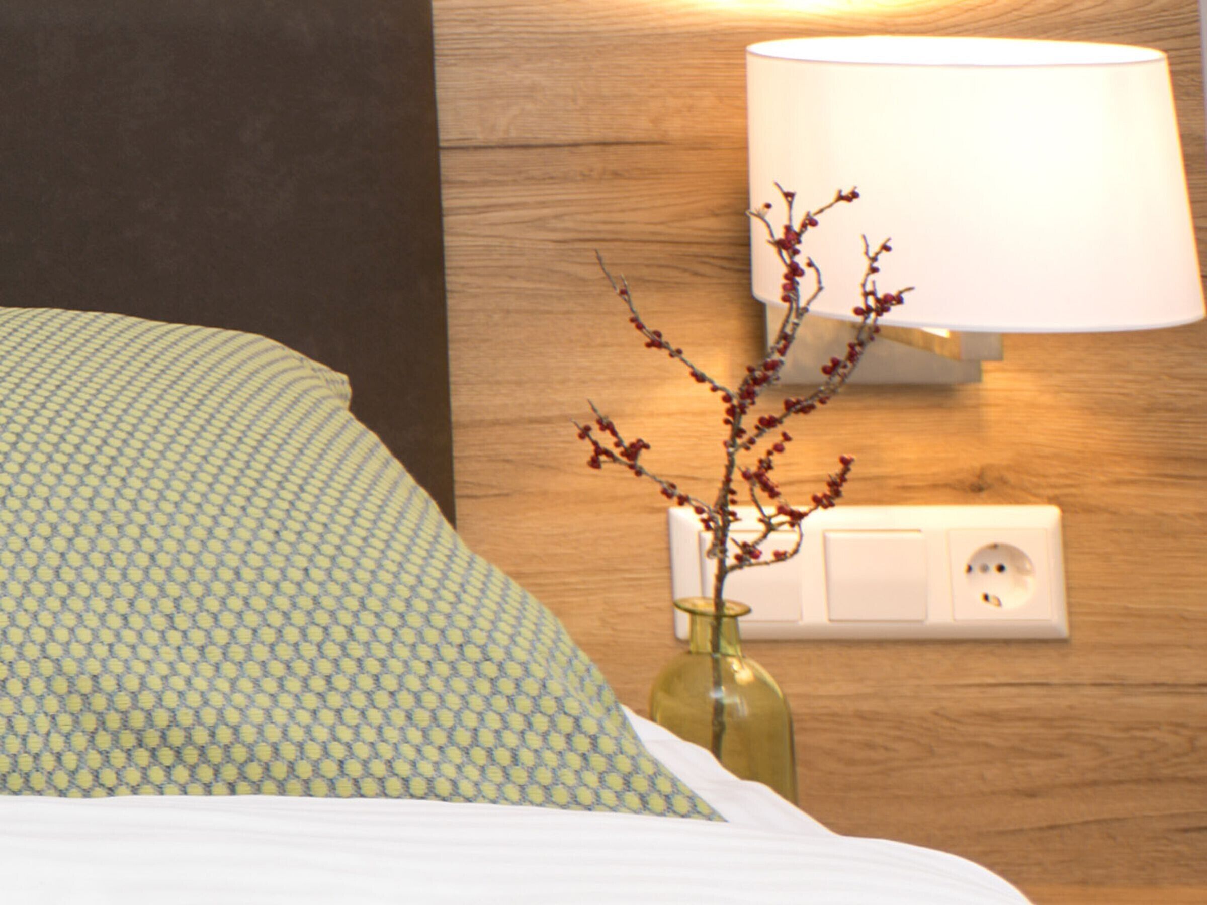 A bed with patterned bedding, a vase with branches, a wall-mounted lamp, and a power outlet.
