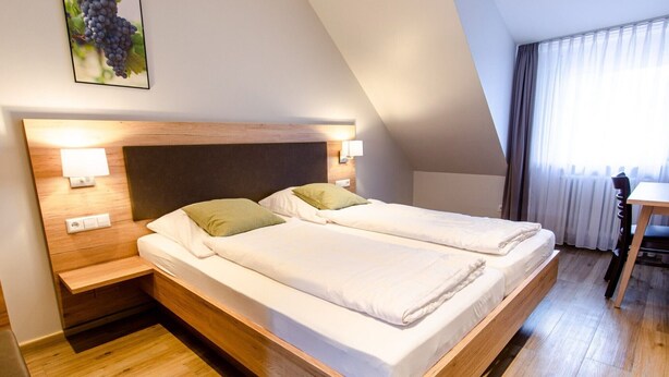 A double bed with a wooden headboard, bedside tables with lamps, and a view of a window with curtains. - Double Room 2 with Balcony and Wi-Fi (Haslach im Kinzigtal)
