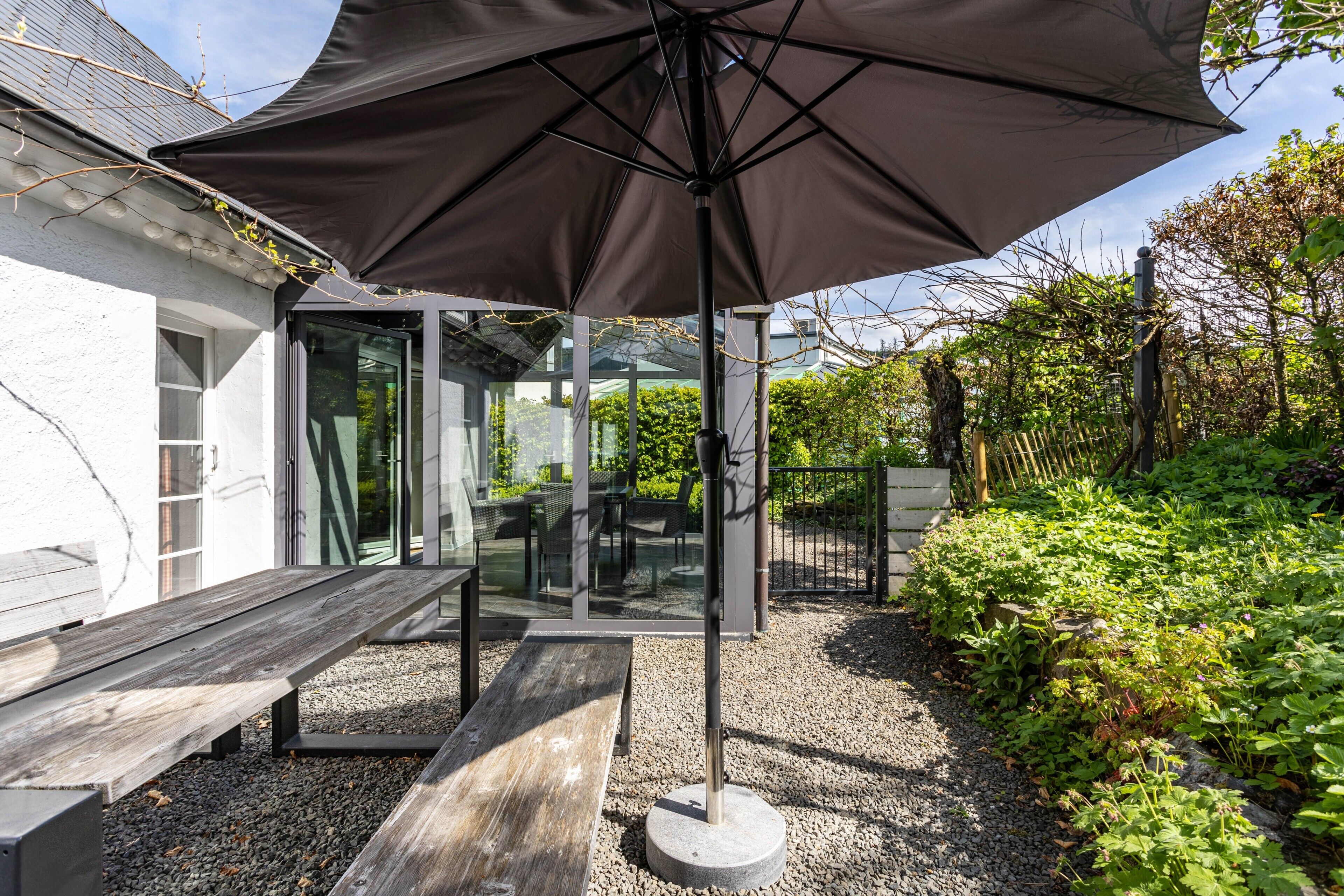 A patio with a table and chairs, a large umbrella, and a garden area with plants and a fence.