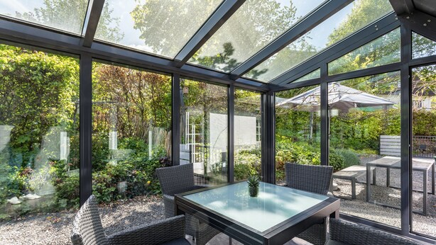Une véranda aux murs de verre, meublée de mobilier en osier, et offrant une vue sur un jardin.