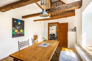 A dining area with a wooden table, chairs, a pendant light, a painting, and a view into another room.