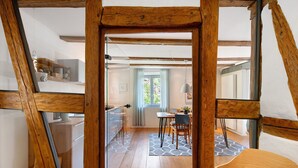 Una habitación con vigas de madera a la vista, una cocina con armarios blancos, una zona de comedor con mesa y sillas, y un ventanal con cortinas.