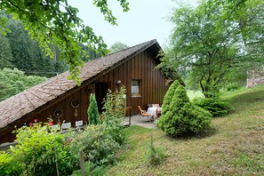 Eine rustikale Holzhütte mit geneigtem Dach, umgeben von üppigem Grün und einem kleinen Garten mit Tisch und Stühlen.