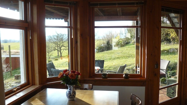 Una mesa de madera para comer con un florero, ventanas amplias con vistas a un paisaje cubierto de césped y una silla.