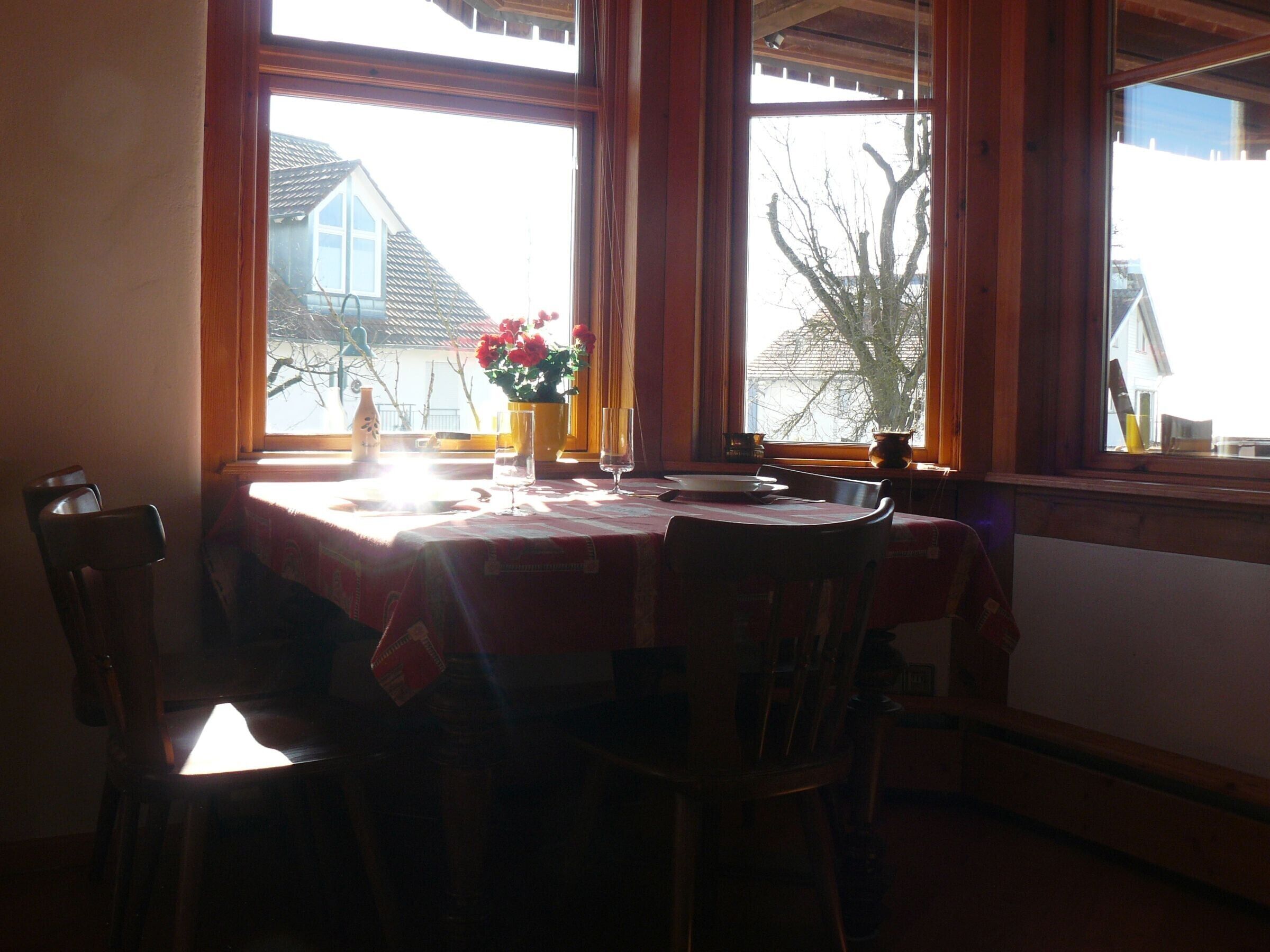 A dining area with a table set for two, a vase with flowers, and a view of a house through large windows.