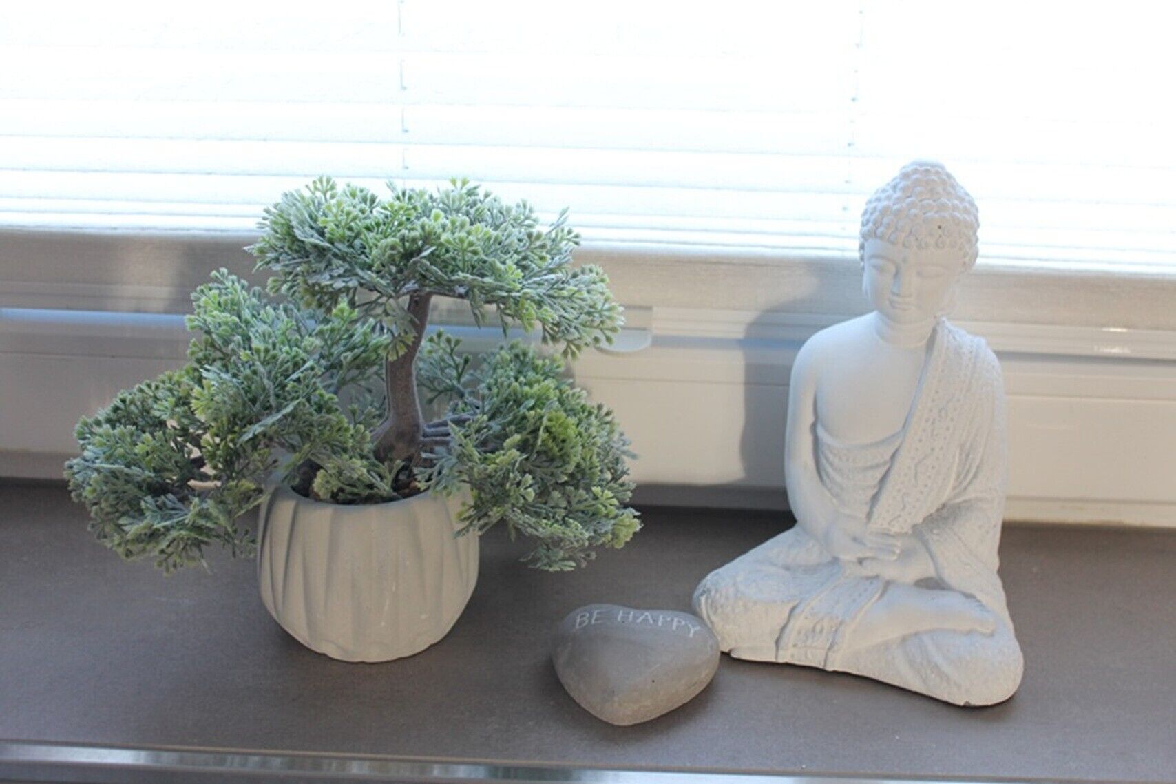 A bonsai tree in a pot, a seated Buddha statue, and a smooth stone with 'BE HAPPY' inscribed on it.