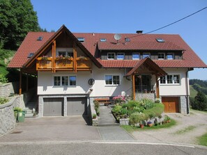 A two-story house with a red-tiled roof, wooden balconies, and a stone wall.