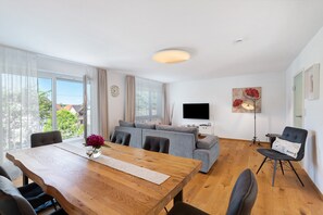 A modern living room with a wooden dining table, grey sofa, and a flat-screen TV.