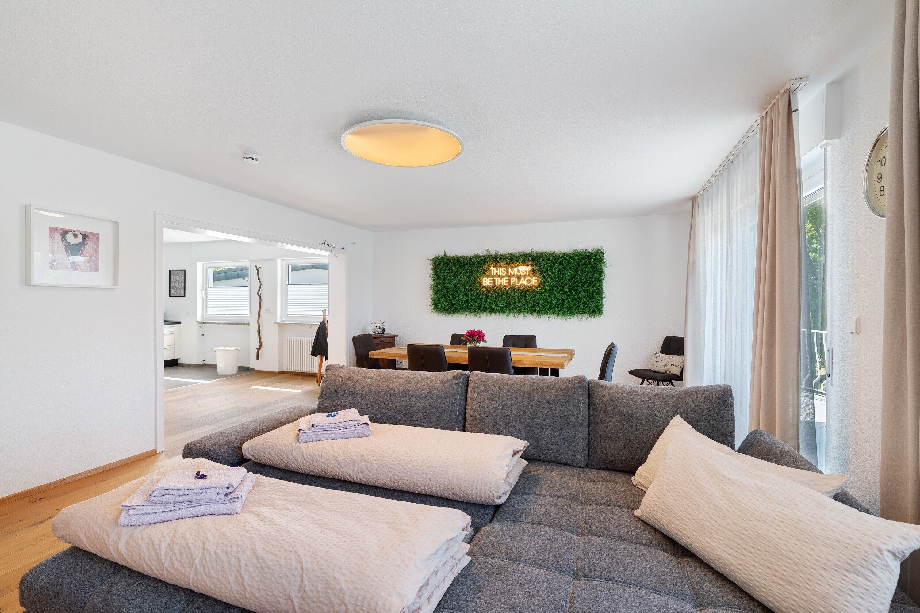 A modern living room with a grey sofa, two beige cushions, a green wall decoration, and a dining area in the background.