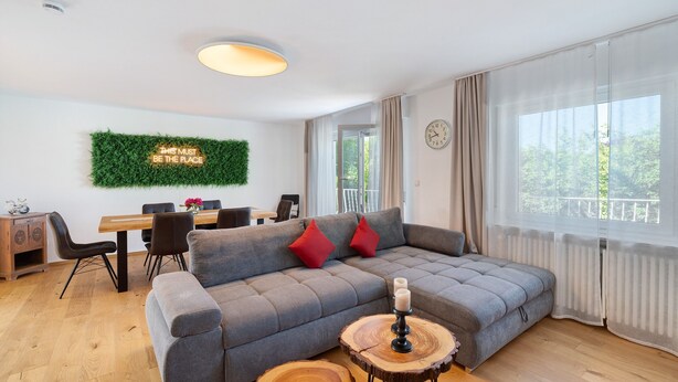 A modern living room with a grey sectional sofa, a dining table, and a large window with curtains. - Apartment 'Weinbergsuite' with Private Terrace, Balcony and Wi-Fi (Kappelrodeck)