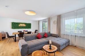 A modern living room with a grey sectional sofa, a dining table, and a large window with curtains.