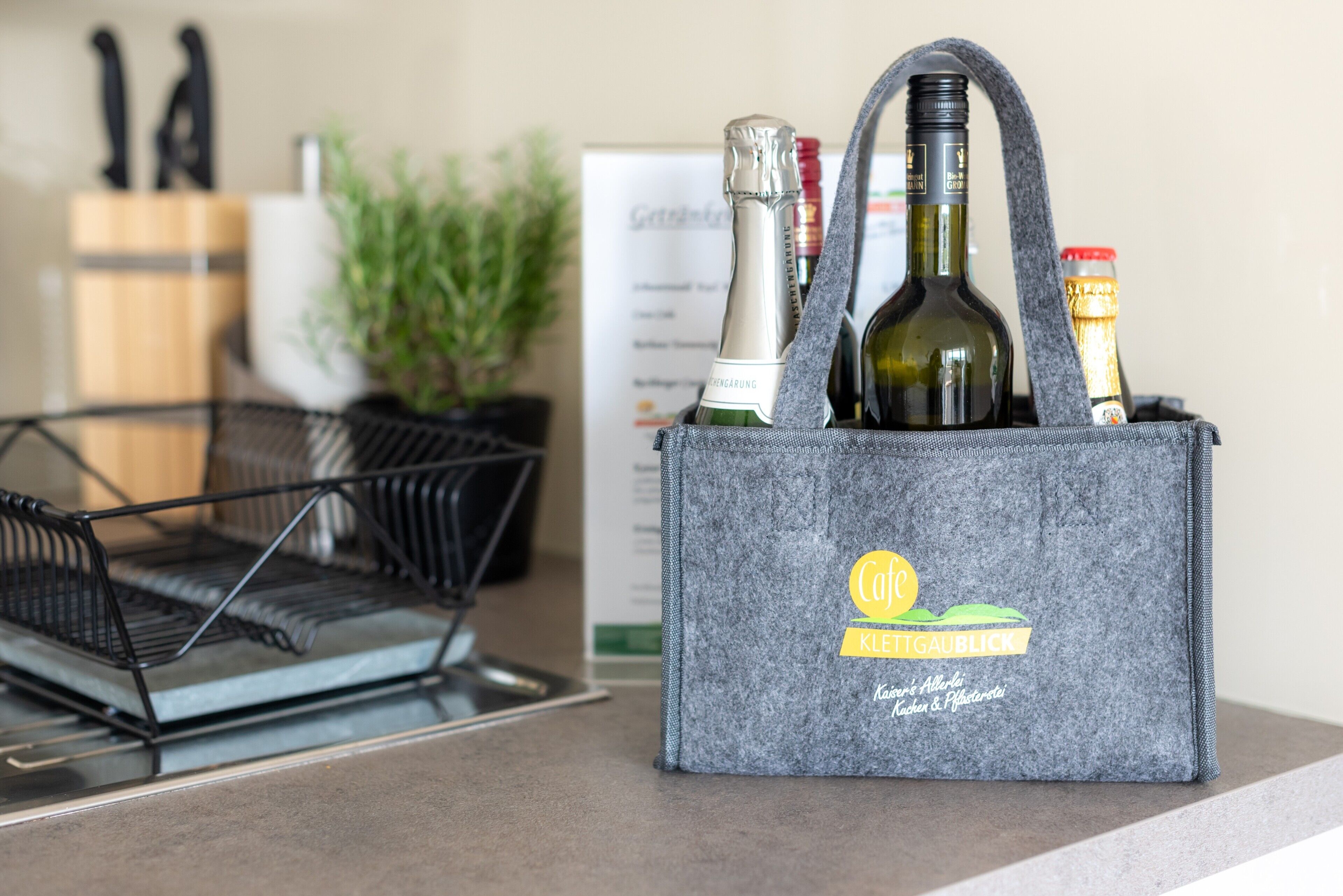 A grey tote bag with a logo, containing bottles of wine and a corkscrew, placed on a kitchen counter.