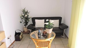 Ein Wohnbereich mit einem schwarzen Sofa, einem Glastisch, einem Korbstuhl, einer Topfpflanze und einem kleinen Tisch mit Flasche und Glas.