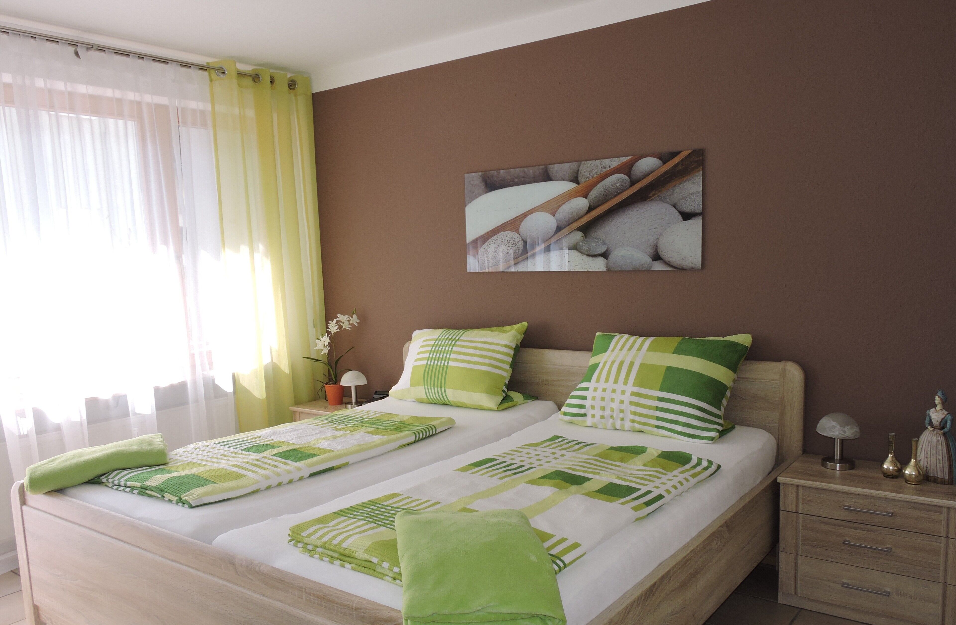A double bed with green and white bedding, a nightstand with a lamp, a painting of stones, and a window with sheer curtains.