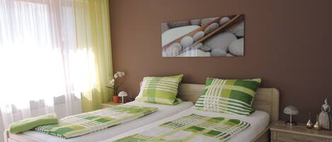 A double bed with green and white bedding, a nightstand with a lamp, a painting of stones, and a window with sheer curtains.
