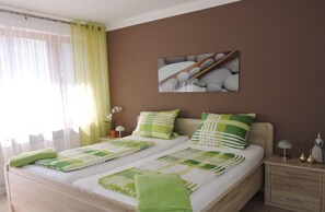 Ein Doppelbett mit grün-weißer Bettwäsche, ein Nachttisch mit Lampe, ein Gemälde mit Steinen und ein Fenster mit durchsichtigen Vorhängen.