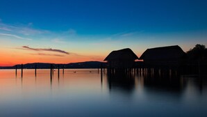 A serene waterfront scene with two thatched-roof structures, likely overwater bungalows, connected by a wooden walkway over calm water.