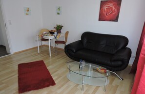 A living room with a black leather sofa, a glass coffee table, a red rug, a dining table with chairs, and a red rose painting on the wall.