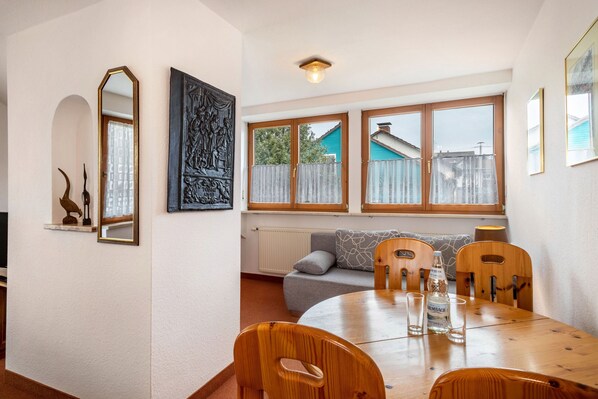 A dining area with a round wooden table, four wooden chairs, a sofa, and a window with a view of a house.