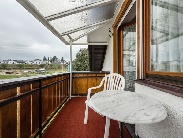 A balcony with a white chair and a small round table, a wooden railing, and a view of houses and greenery.