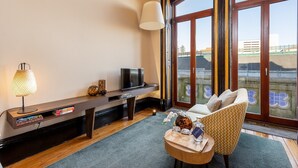 Een moderne woonkamer met een televisie, boeken, een lamp en uitzicht op een gebouw door grote ramen.